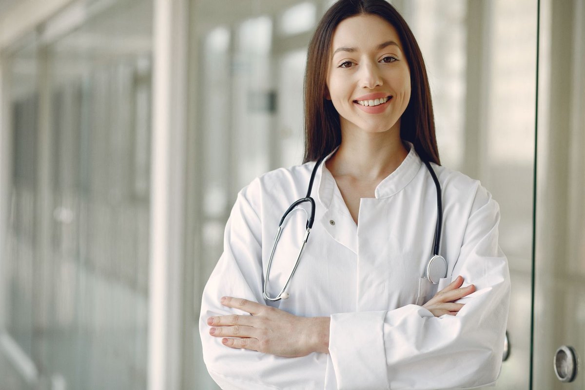 Smiling doctor in a white coat with a stethoscope, arms crossed, in a bright hospital corridor